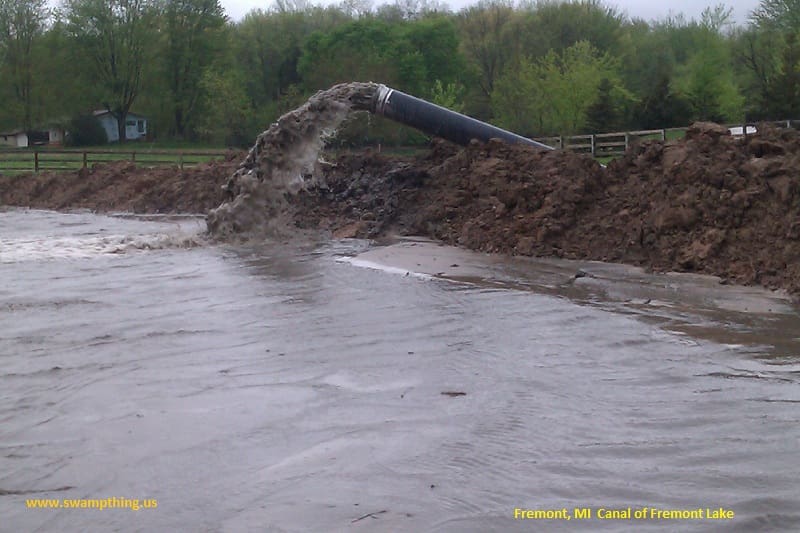 A large pipe is in the mud near some trees.